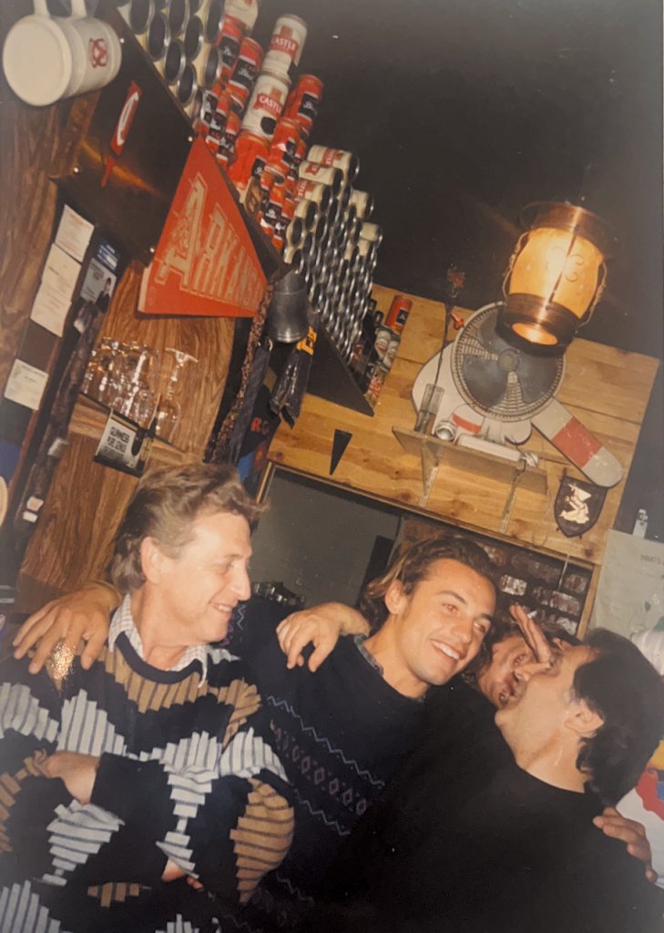 Andrea abbracciato agli amici al bancone di Buddy's, sotto la mensola piena di lattine di Castle