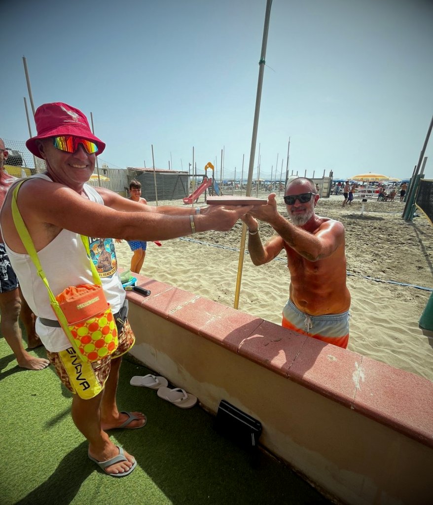 Lepre consegna il Tagliere di Legno a Buoso sopra il muretto della spiaggia, con i campi da beach tennis sullo sfondo